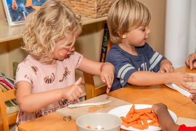 Two preschoolers practicing food prep by cutting carrots at a wooden table